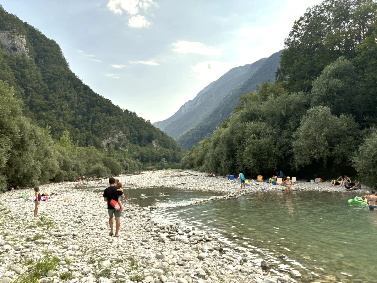 People enjoying Nadiža river and sitting on the pebble and rock riverbanks with green trees and hills in the background